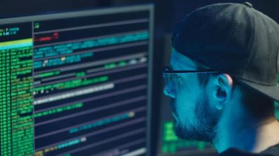 Perfil de una persona con gorra y gafas analizando líneas de código y datos en un monitor en un entorno oscuro.