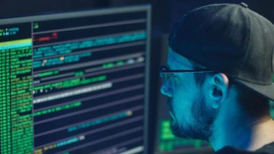 Perfil de una persona con gorra y gafas analizando líneas de código y datos en un monitor en un entorno oscuro.