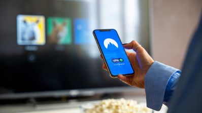 Ilustración del uso de una VPN en el salón de casa, con una persona configurando la conexión en su móvil para proteger el streaming en una Smart TV.