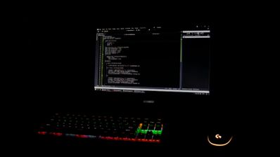 Pantalla de ordenador con líneas de código en un entorno oscuro y teclado RGB iluminado.