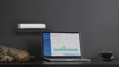 Router profesional QNAP modelo QHora-301W en un entorno de oficina, con un portátil mostrando la nueva interfaz del Sistema de Prevención de Intrusiones (IPS) del firmware v2.6.0.