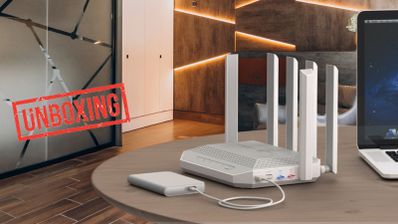 Router inalámbrico Keenetic Hero 5G de color gris con cuatro antenas externas sobre una mesa de madera junto a un disco duro portátil y un portátil.