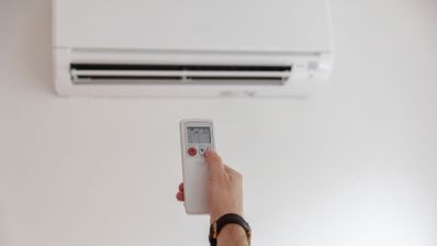 Mano accionando el mando a distancia hacia una unidad de aire acondicionado tipo split instalada en una pared blanca.