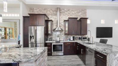 Cocina moderna con armarios de madera oscura, encimeras de granito veteado y electrodomésticos de acero inoxidable bajo una campana extractora sobre pared de piedra.