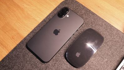iPhone de color oscuro junto a un Magic Mouse sobre una alfombrilla gris en una mesa de madera.