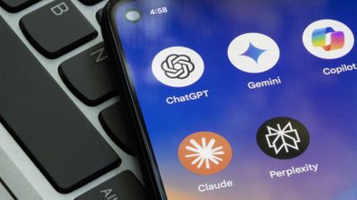 Primer plano de un teléfono móvil sobre el teclado de un portátil mostrando iconos de aplicaciones de inteligencia artificial como ChatGPT, Gemini, Copilot, Claude y Perplexity.
