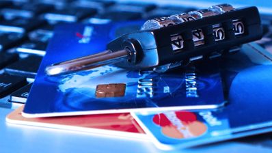 Primer plano de un candado de combinación negro sobre varias tarjetas bancarias situadas encima del teclado de un ordenador portátil con luz azulada.