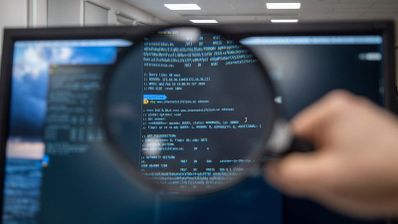 Primer plano de una lupa sostenida frente a una pantalla de ordenador que muestra líneas de código de programación en una terminal.