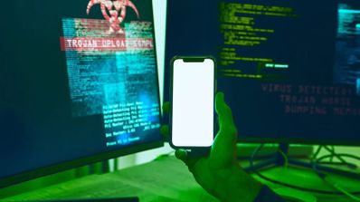 Mano sujetando un teléfono móvil frente a dos monitores que muestran líneas de código y mensajes de advertencia de virus y troyanos en color verde y rojo.