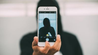Persona con sudadera oscura sosteniendo un teléfono móvil blanco para hacerse un autorretrato o grabar un vídeo selfie.