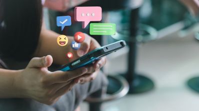 Primer plano de manos sosteniendo un smartphone del que emergen iconos 3D de notificaciones, mensajes y reacciones de redes sociales.