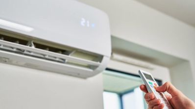 Mano sujetando un mando a distancia frente a un aparato de aire acondicionado tipo split configurado a 21 grados.