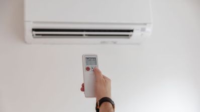 Mano de una persona ajustando la temperatura con un mando a distancia frente a un aparato de aire acondicionado tipo split de color blanco.
