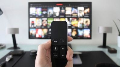 Primer plano de una mano sujetando un mando a distancia frente a una televisión con un catálogo de aplicaciones de streaming.