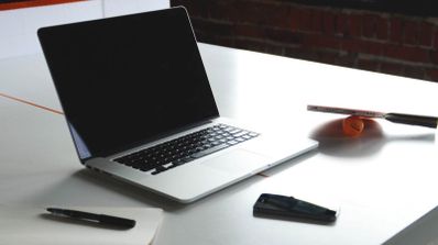 Ordenador portátil abierto sobre una mesa escritorio blanca junto a una libreta, un bolígrafo y una pala de ping-pong al fondo.