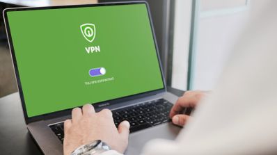 Manos de una persona utilizando un ordenador portátil que muestra en pantalla un software de conexión VPN activa con fondo verde y un icono de escudo.