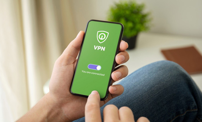 Mano sosteniendo un smartphone con una aplicación de VPN activada sobre un fondo verde con icono de escudo.