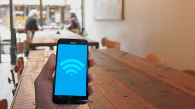 Mano sosteniendo un teléfono móvil con el icono de conexión Wi-Fi en pantalla azul dentro de una cafetería.