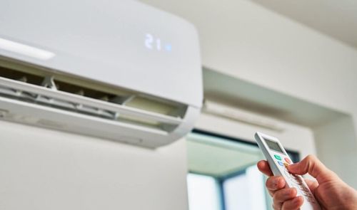 Mano utilizando un mando a distancia frente a un aparato de aire acondicionado tipo split configurado a 21 grados.