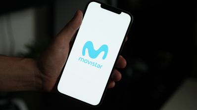 Mano sujetando un teléfono móvil que muestra el logotipo azul de Movistar sobre un fondo blanco.