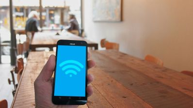 Mano sujetando un teléfono móvil que muestra el símbolo de conexión Wi-Fi en pantalla sobre el fondo desenfocado de una cafetería.