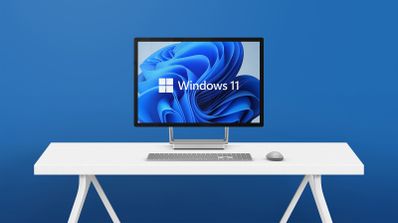 Monitor de ordenador en un escritorio blanco mostrando el logotipo y fondo de pantalla de Windows 11 sobre un fondo azul.
