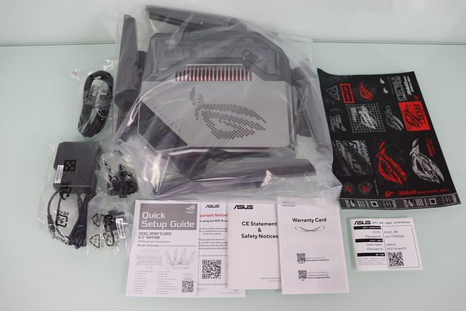 Contenido completo de la caja del router ASUS GT-BN98 sobre una mesa blanca, incluyendo cables, fuente de alimentación y manuales.