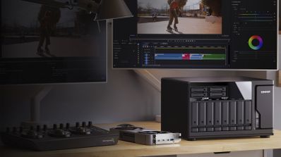 Servidor NAS QNAP TVS-h1277AX en un escritorio junto a monitores con software de edición de vídeo.