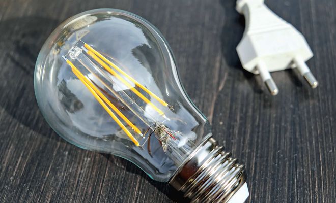 Bombilla eléctrica de filamento LED apagada junto a un enchufe blanco sobre fondo oscuro.