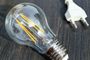 Bombilla eléctrica de filamento LED apagada junto a un enchufe blanco sobre fondo oscuro.