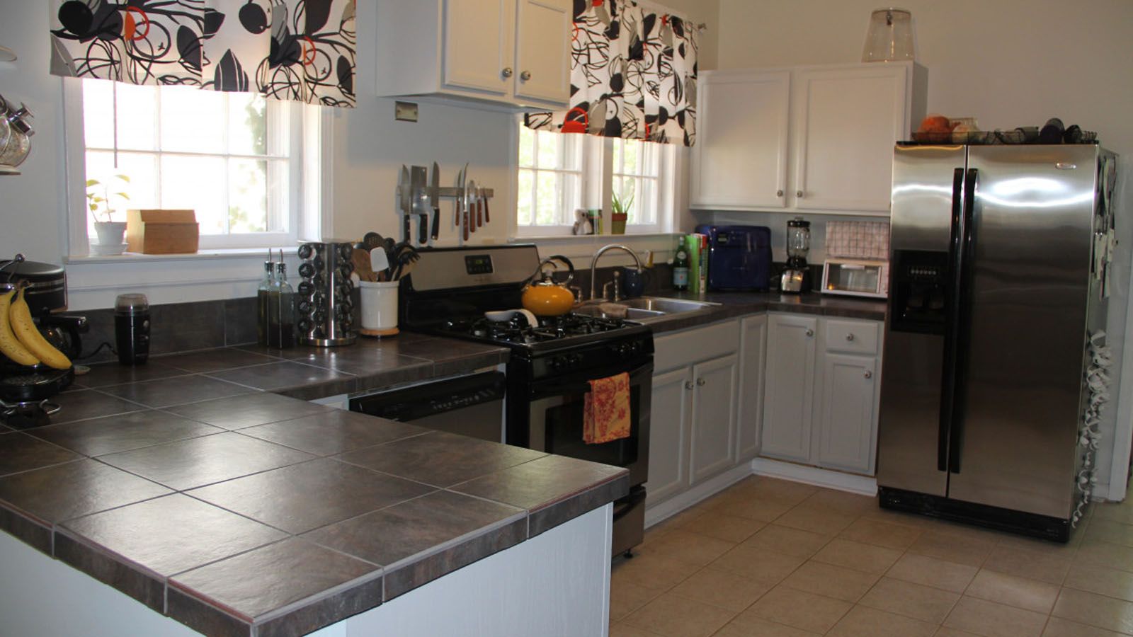 Interior de una cocina doméstica con encimera de baldosas oscuras, armarios blancos y electrodomésticos de acero inoxidable.