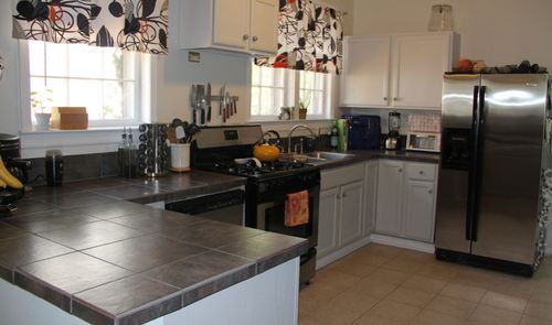 Interior de una cocina doméstica con encimera de baldosas oscuras, armarios blancos y electrodomésticos de acero inoxidable.