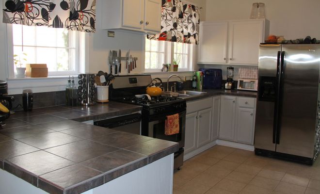 Interior de una cocina doméstica con encimera de baldosas oscuras, armarios blancos y electrodomésticos de acero inoxidable.