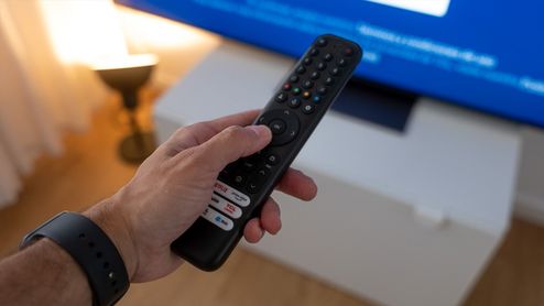 Mano de una persona sujetando un mando a distancia frente a una televisión encendida.