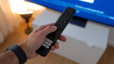 Mano de una persona sujetando un mando a distancia frente a una televisión encendida.