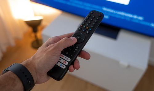 Mano de una persona sujetando un mando a distancia frente a una televisión encendida.