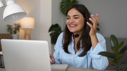 Mujer sonriente con auriculares y micrófono hablando frente a su ordenador portátil durante una videollamada.