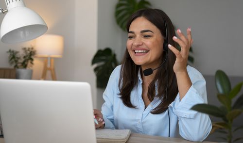 Mujer sonriente con auriculares y micrófono hablando frente a su ordenador portátil durante una videollamada.