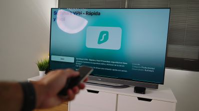 Mano sosteniendo un mando a distancia frente a un televisor que muestra la interfaz de la aplicación Surfshark VPN.