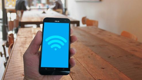 Mano sosteniendo un teléfono móvil que muestra el icono de conexión Wi-Fi en pantalla sobre un fondo de mesas de madera.