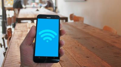 Mano sosteniendo un teléfono móvil que muestra el icono de conexión Wi-Fi en pantalla sobre un fondo de mesas de madera.
