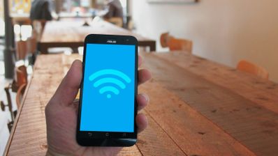 Mano sosteniendo un teléfono móvil que muestra el icono de conexión Wi-Fi en pantalla sobre un fondo de mesas de madera.