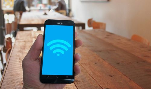 Mano sosteniendo un teléfono móvil que muestra el icono de conexión Wi-Fi en pantalla sobre un fondo de mesas de madera.