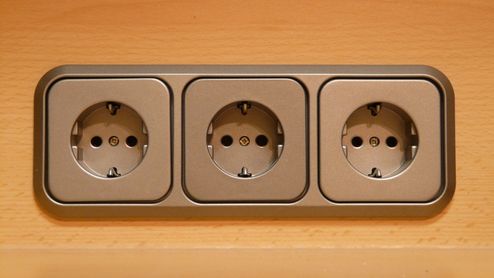 Tres tomas de corriente Schuko integradas en un marco triple de color gris sobre una superficie de madera.