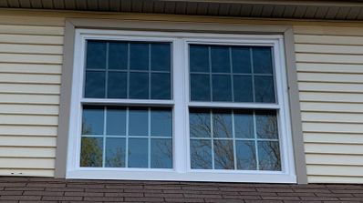 Ventana blanca de doble hoja con cuadrícula en la fachada de una vivienda con revestimiento exterior beige.
