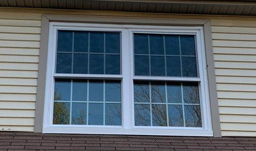 Ventana blanca de doble hoja con cuadrícula en la fachada de una vivienda con revestimiento exterior beige.