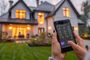 Manos sujetando un teléfono móvil con una aplicación de control domótico frente a una vivienda unifamiliar iluminada al atardecer.
