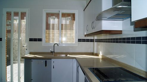 Cocina doméstica con muebles blancos, encimera de madera clara y ventana sobre el fregadero.