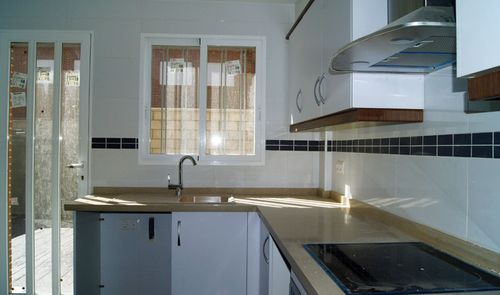 Cocina doméstica con muebles blancos, encimera de madera clara y ventana sobre el fregadero.