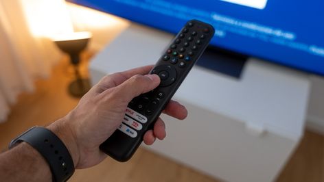 Primer plano de una mano sosteniendo un mando a distancia frente a una Smart TV con pantalla azul.